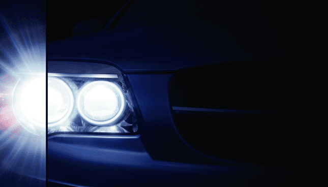 Un primer plano de la luz frontal de la parte izquierda de un coche iluminando la oscuridad. Una línea vertical divide la luz en dos, con efecto destello de la luz en la parte izquierda de la línea.