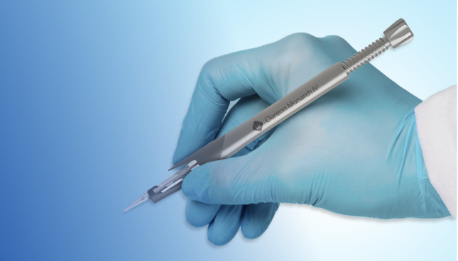 Una imagen de sistema inyector Clareon Monarch IV sujetado por una mano con un guante quirúrgico azul. La mano agarra el dispositivo como agarramos un bolígrafo y aparece sobre un fondo azul.