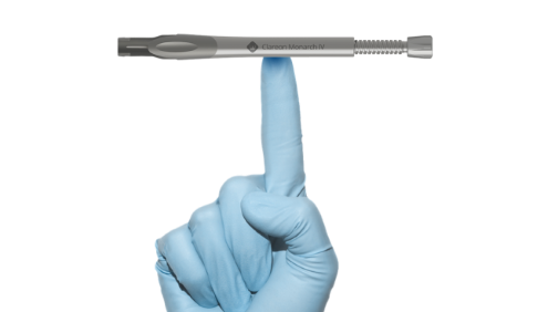 Hand wearing a disposable glove with the index finger pointing upwards. The Clareon Monarch IV Handpiece sits horizontally, balanced on top of the index finger.