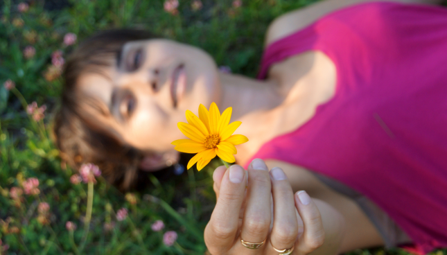 Woman laying in the grass, holding up a yellow flower
